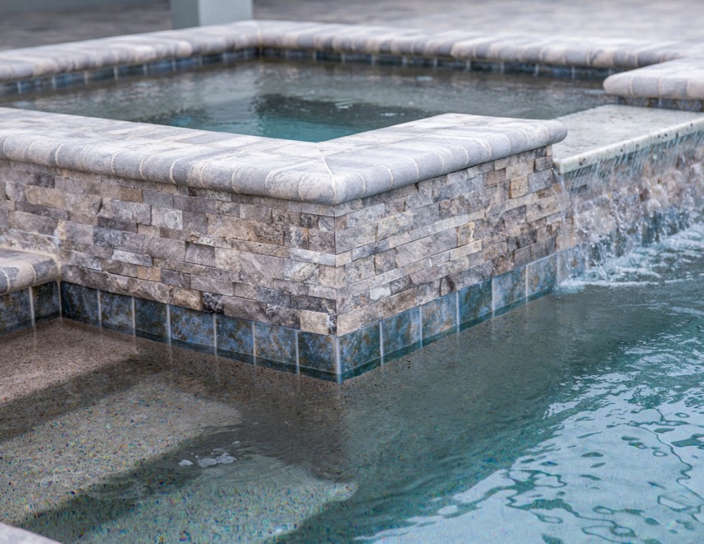 Raised spa with stone finish and spillway flowing into a swimming pool